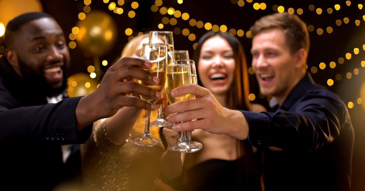 A group of friends gathered around smiling and clinking their glasses together underneath some fairy lights.
