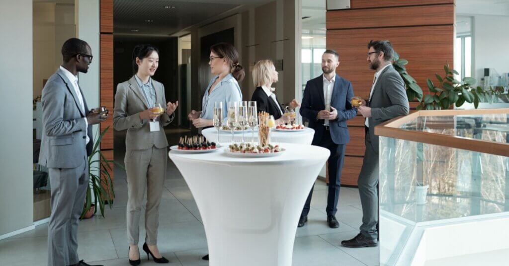 A group of professionals in suits standing around some tables. They are holding drinks and talking to each other.