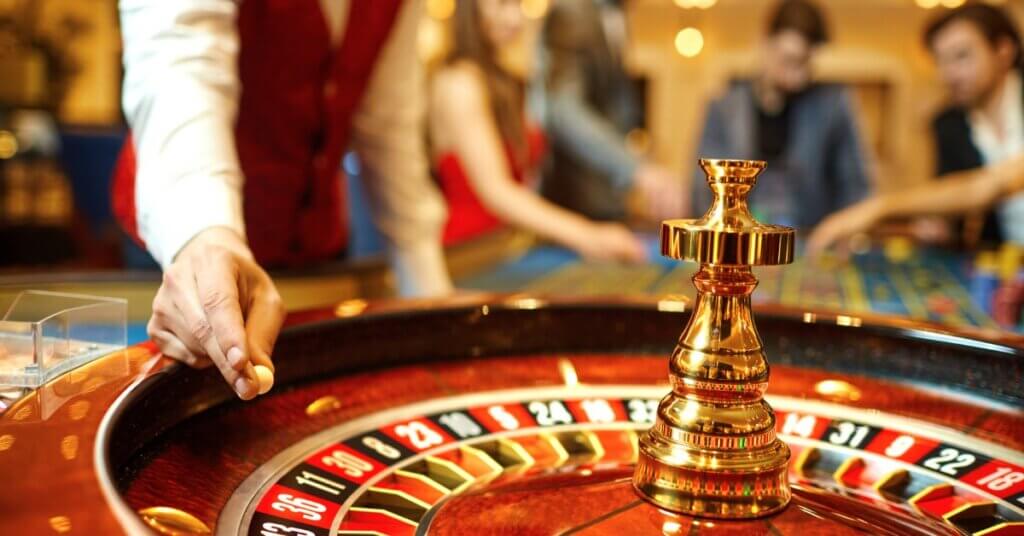 A roulette dealer holding the roulette ball as he's about to drop it into the wheel. A group of people wait behind him.