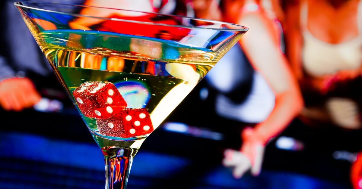 A closeup on a martini glass full of a slightly yellow liquid. Two red dice are floating around in the liquid.