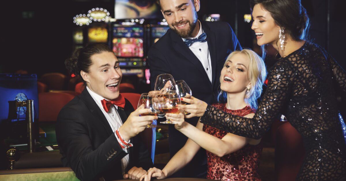 Two men and two women dressed in fancy clothes tapping their glasses together above a roulette table.