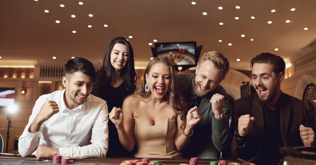 A group of five happy people looking at their winnings in poker chips that are on top of a roulette table.