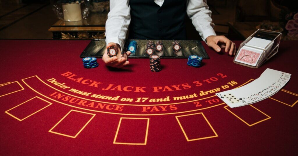 A blackjack dealer standing behind a blackjack table with a poker chip in his hand. More poker chips are in front of him.