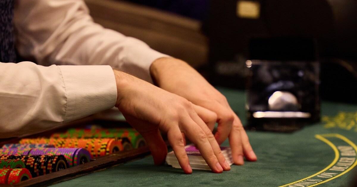 A blackjack dealer placing a deck of cards down onto a blackjack table in front of a bunch of casino chips.